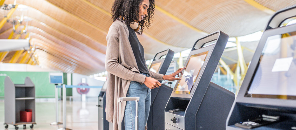 Traveler using a touchscreen self-service kiosk at an airport check-in or bag drop area, with luggage beside her